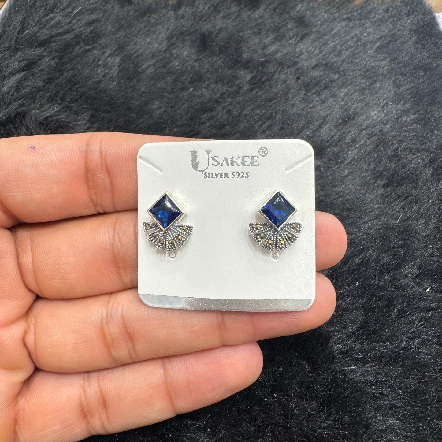 Pair of silver earrings with blue gemstones on a U.SAKKE branded card held by a hand against a dark textured background.